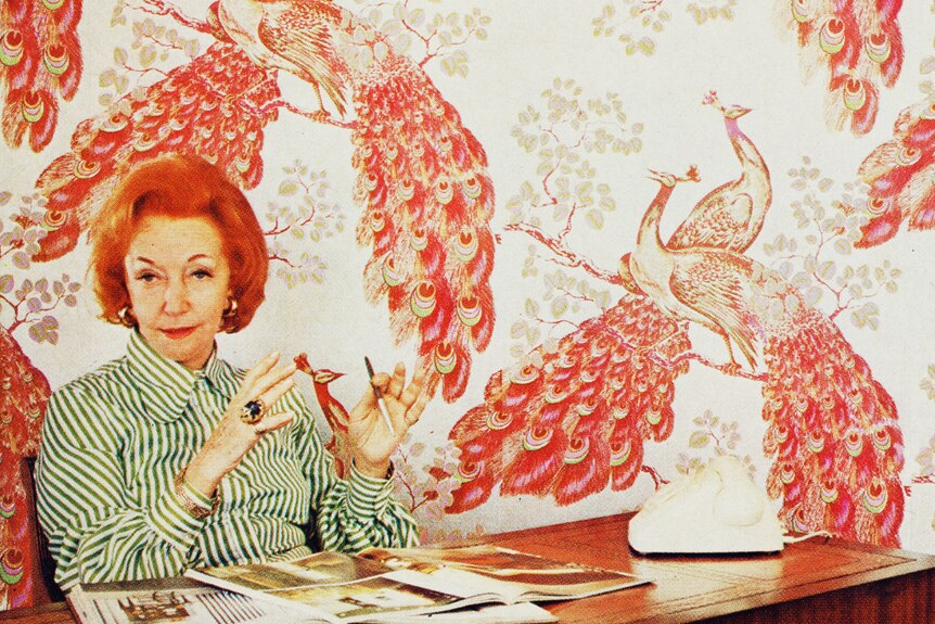 A woman with short red hair sits behind a desk, with books on it, in front of peacock-prinyed wallpaper. 