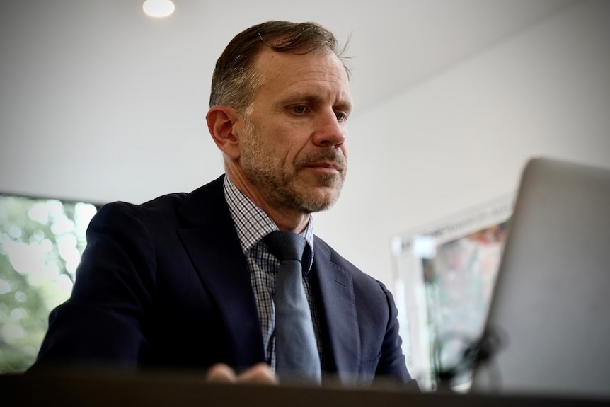 A middle-aged man wearing a navy suit and tie sits looking at his laptop.