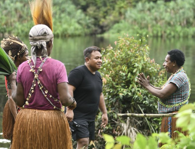 A bulky man in a black t-shirt is surrounded by three indigenous people.
