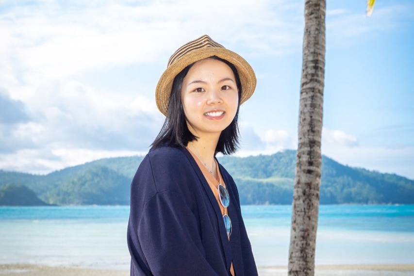 a woman wearing a sun hat smiles at the camera, there is a beach behind her.