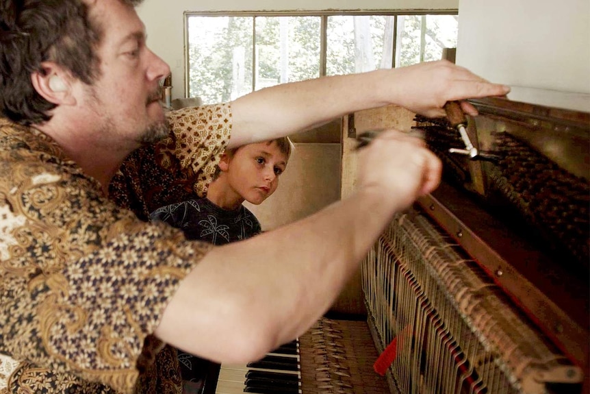 man busy tuning a piano with a tuning tool in a home while a young boy is watching