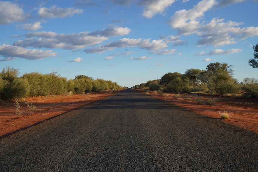 Paved road with red dirt either side, blue sky with clouds above