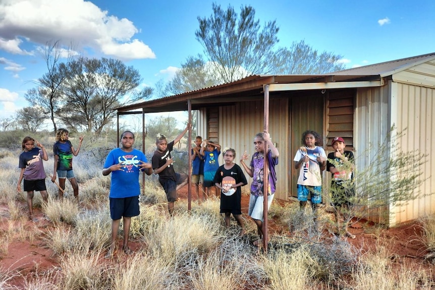 10 Indigenous children stand in the bush in red dirt outside a metal shed. 
