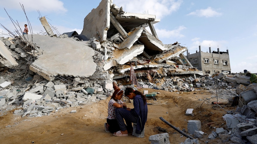 a massive rubble of a house is behind two little girls crouching and talking