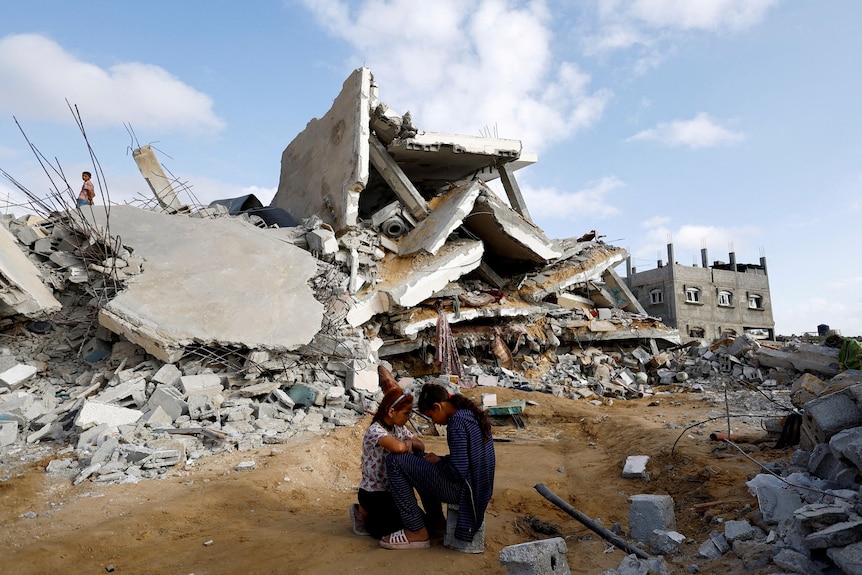 a massive rubble of a house is behind two little girls crouching and talking
