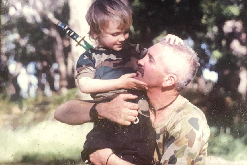 A young Zaynn Bird in a camouflage print t-shirt smiles at his dad mid-hug outside on the lawn.