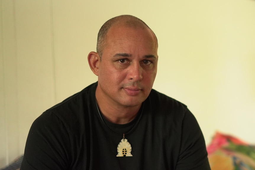 Man with black shirt and necklace sits on couch behind yellow wall