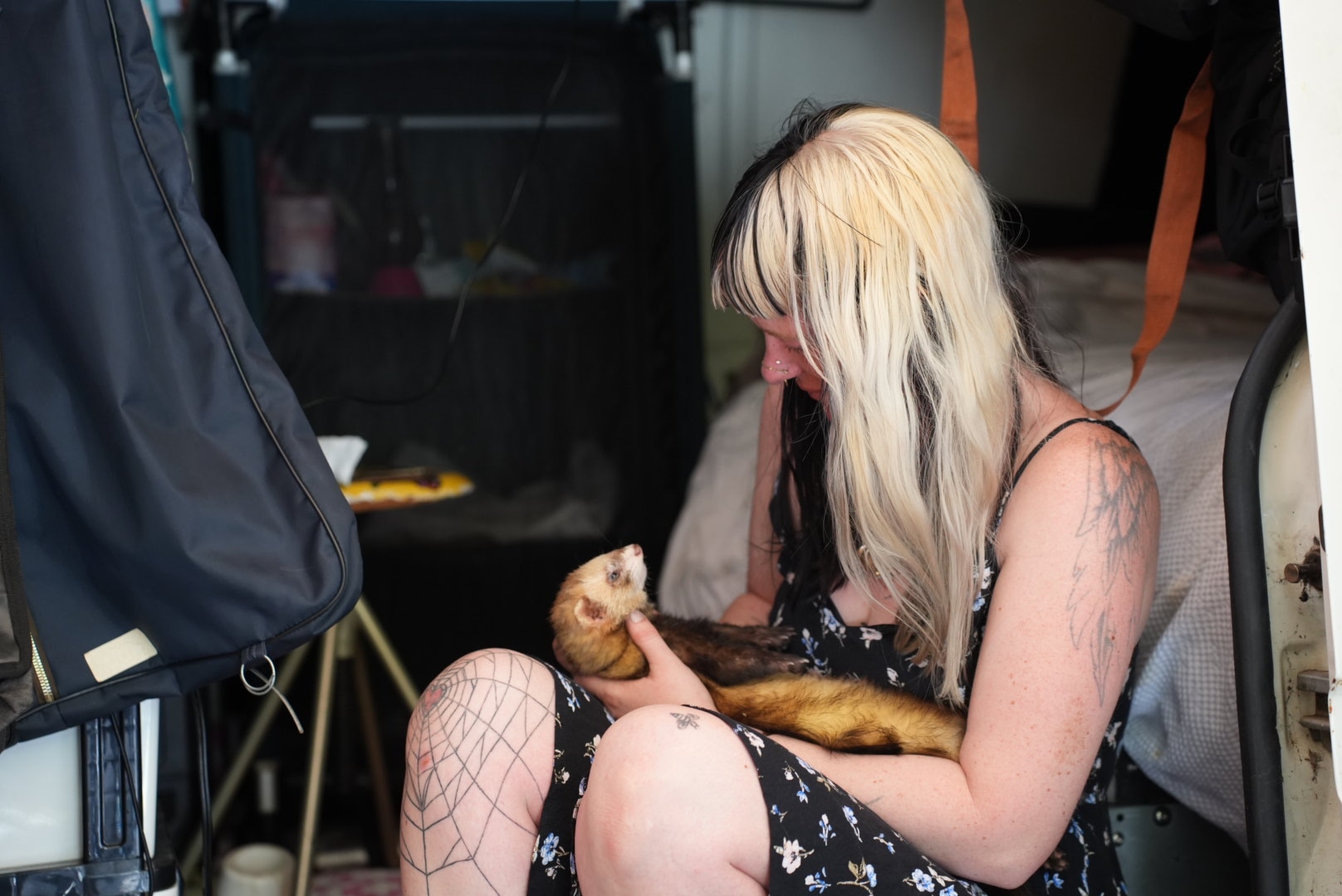 A woman with blond hair and tattoos on her arms and legs plays with a pet ferret in a van