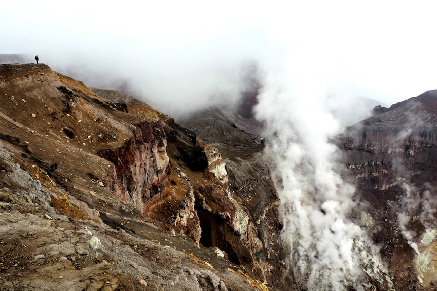 A man stands on a cliff top above a deep crater with steam rising from the middle.