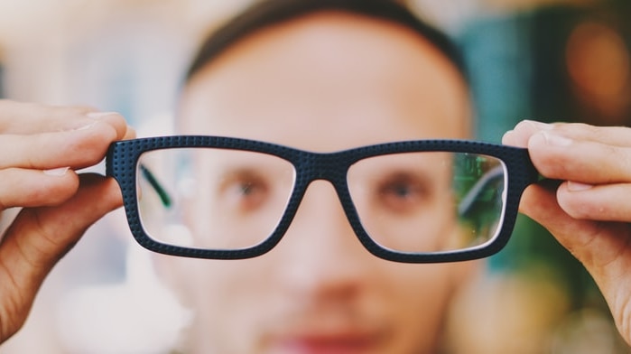 Man holds glasses in front of him. His glasses and hands are in focus his face behind them is fuzzy.