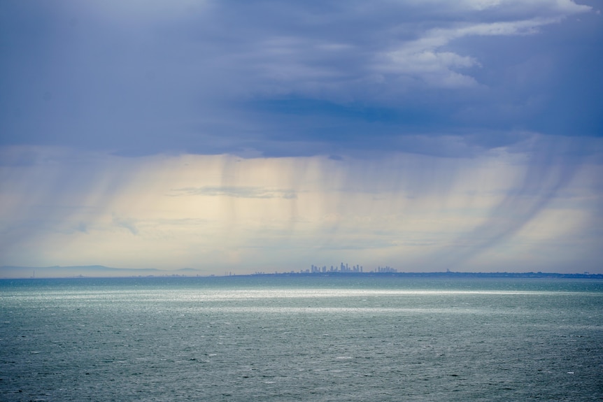 Clouds and rainfall build up over a long shot of city of Melbourne, ocean in foreground.
