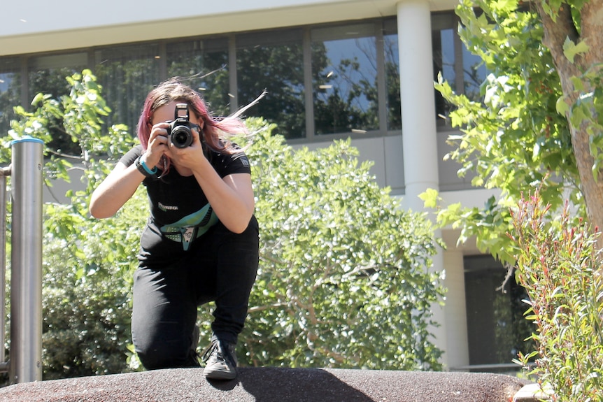 A young woman with pink hair holds a camera up as she crouches on a fence to take a photo.