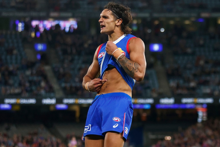 A Western Bulldogs AFL player lifts his jersey and points to his stomach during a match against the Brisbane Lions.
