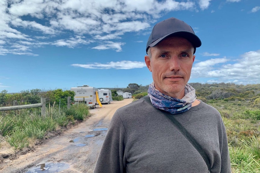 A man wearing a cap stands on a dirt road with several caravans behind him.