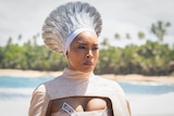 Black woman wearing silver headdress that fans out in a cone shape and patterned white and cream cape stands on a shore edge.