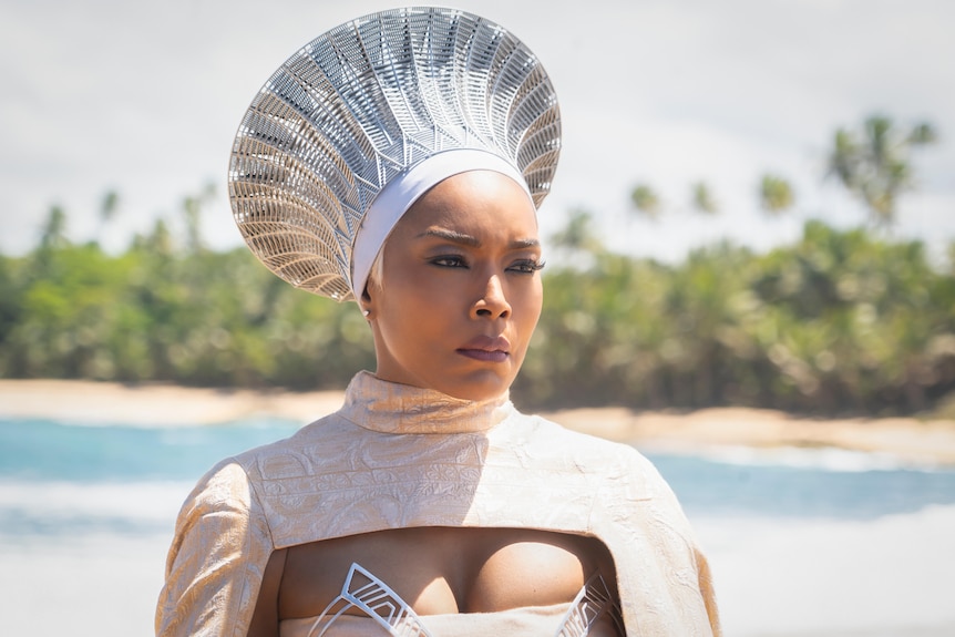 Una mujer negra que lleva un tocado plateado en forma de cono y un tocado estampado en blanco y crema se encuentra al borde de la playa.