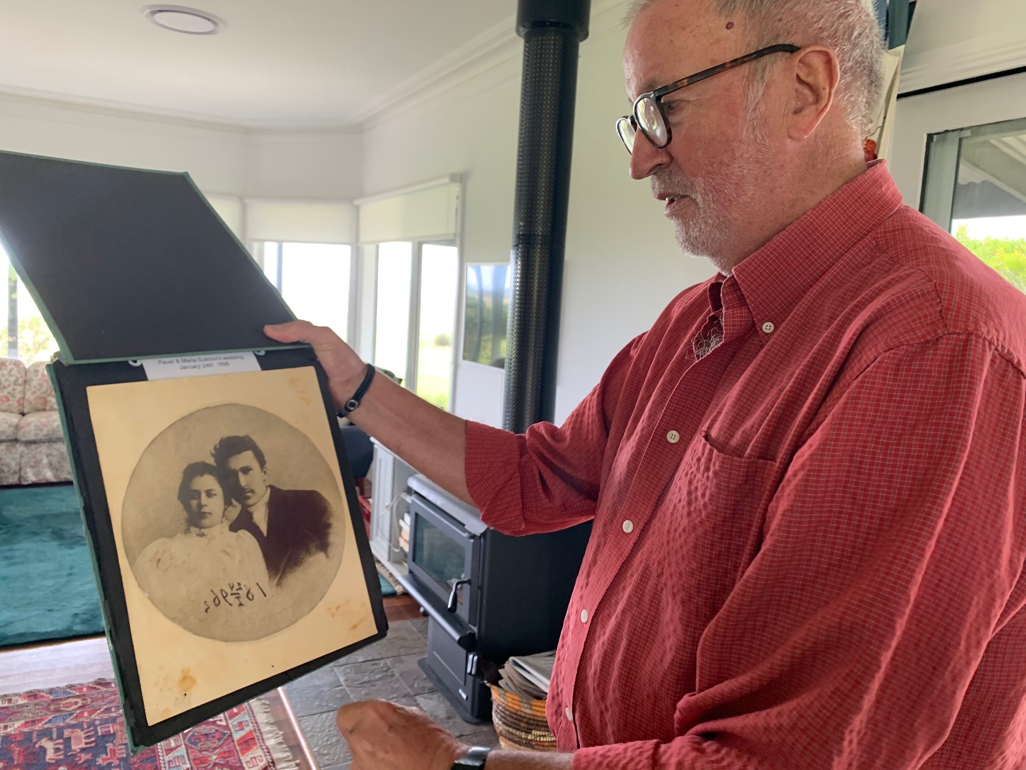 Man in red shirt holds an old photo in a black frame at arms length and appears to study it.