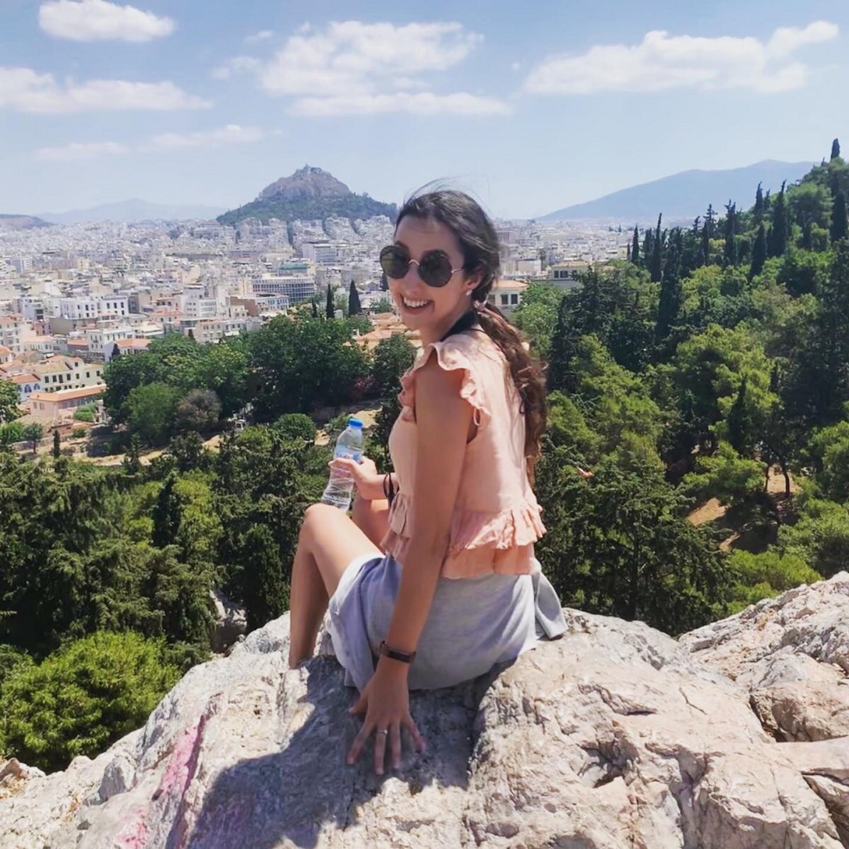 Inez sitting on a rock overlooking stunning buildings and mountains, smiling at the camera.