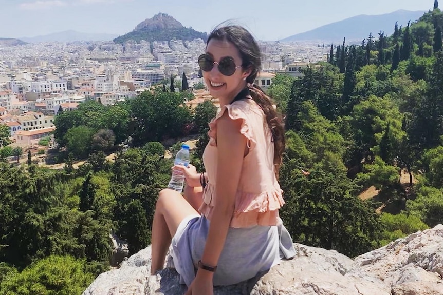 Inez sitting on a rock overlooking stunning buildings and mountains, smiling at the camera.