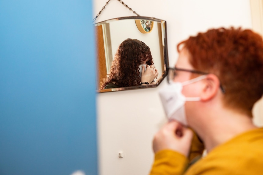 A brunette and redhead woman puts on her mask in front of a mirror