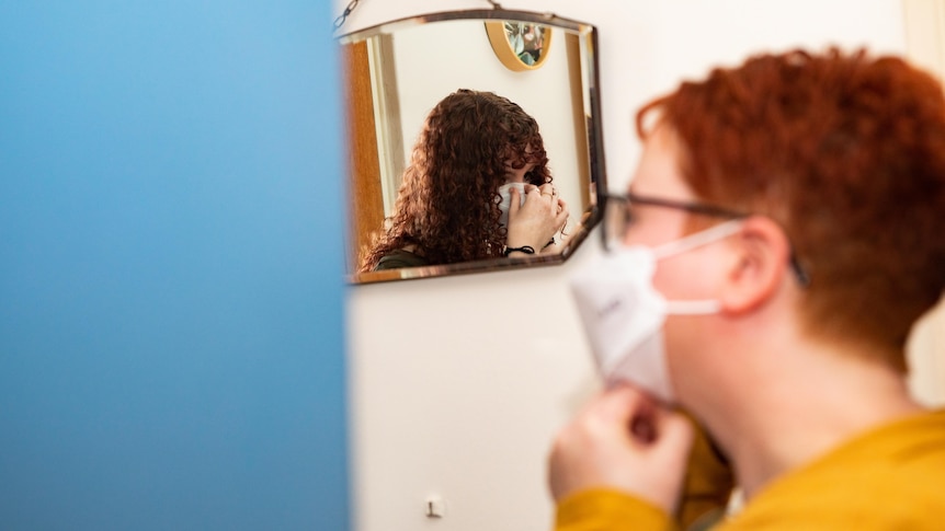 A brunette and red haired lady put on their masks in front of a mirror