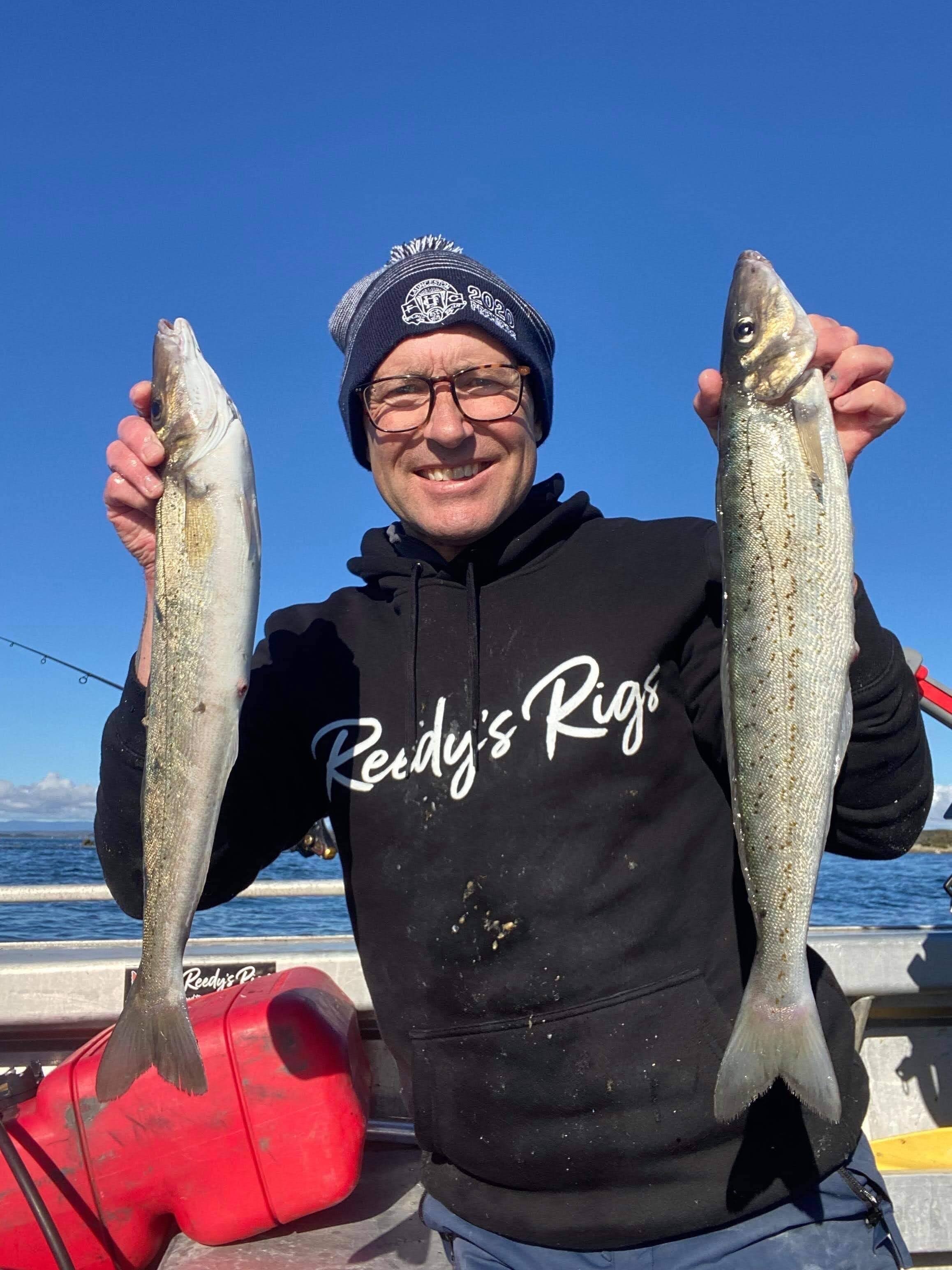 A man in a beanie holds up two long fish he has caught.