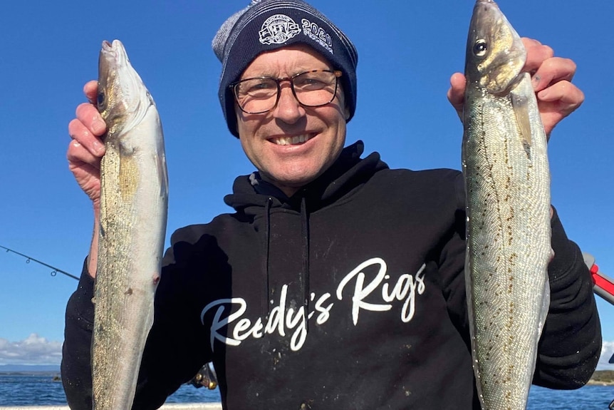 A man in a beanie holds up two long fish he has caught.