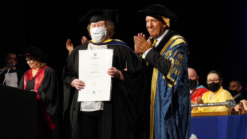A woman with Down syndrome wearing an academic gown holds a certificate with a man clapping beside her