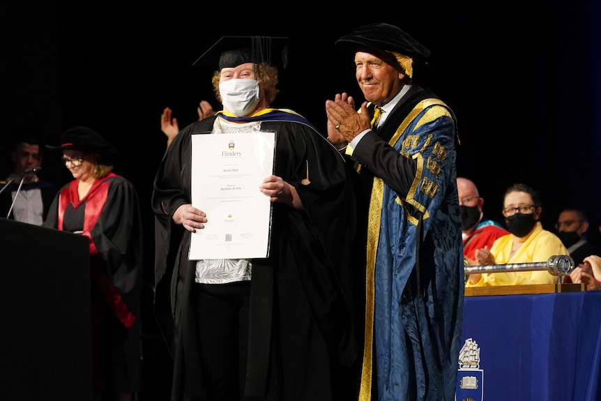 A woman with Down syndrome wearing an academic gown holds a certificate with a man clapping beside her