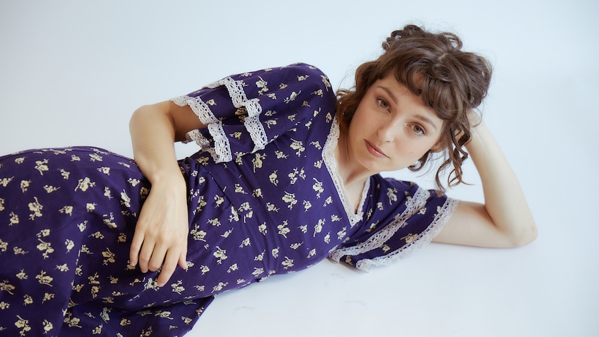 A 2022 press shot of Stella Donnelly in a blue frock lying down on her side - elbow propping up head