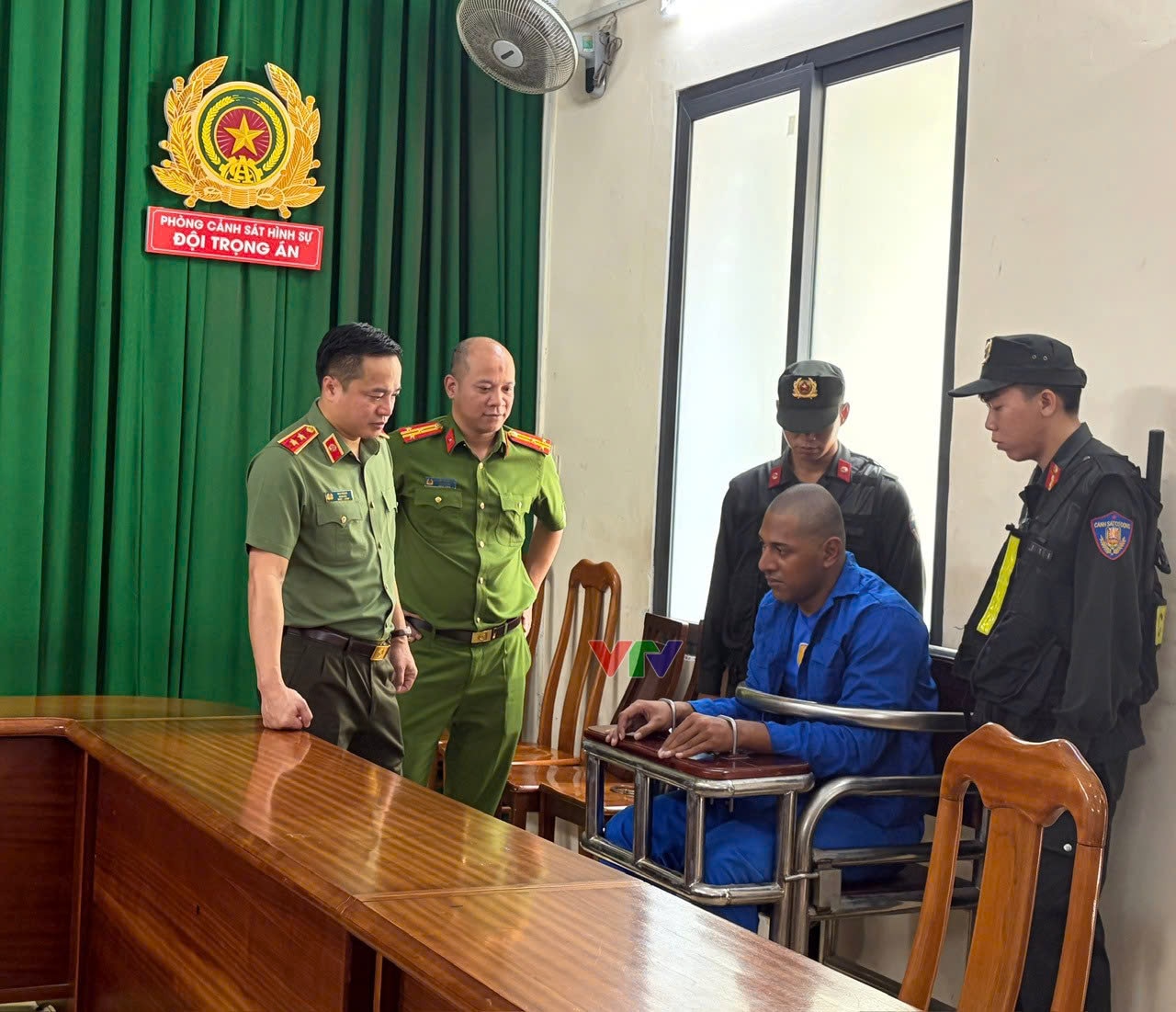 Vaa Vaa 坐在椅子上，双手被绑在旁边的小桌子上，腰间系着一根金属杆，周围都是警察。