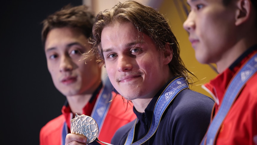 An Australian diver looks at the camera while holding a gold medal, as two other medallists stand next to him.