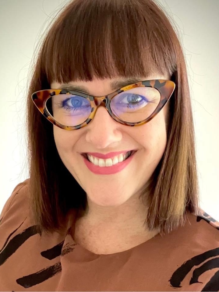 a lady with reddish, brown hair and wide brimmed glasses smiles at the camera