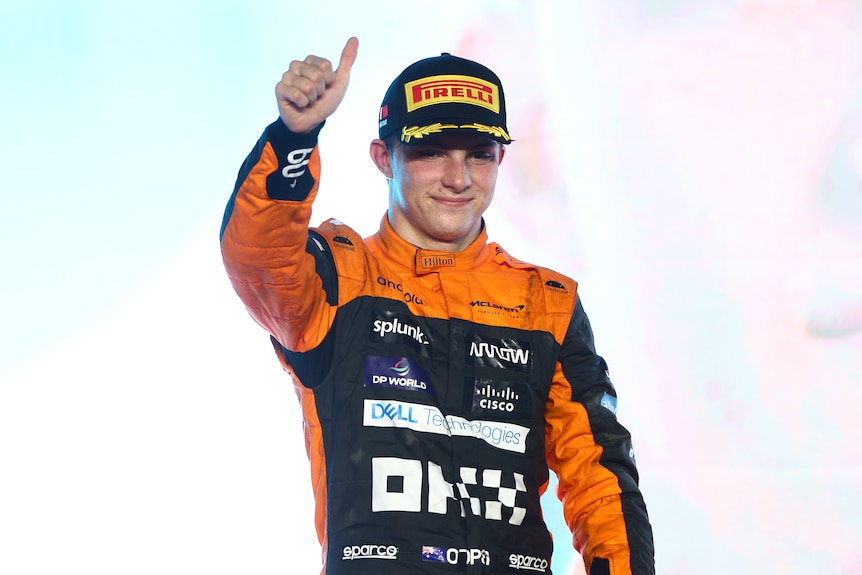 An F1 driver in an orange race suit, black cap, gives a thumbs-up on the podium after a race.