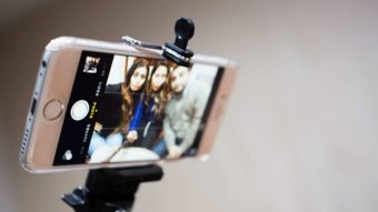 Three people look at an iphone in a selfie stick.