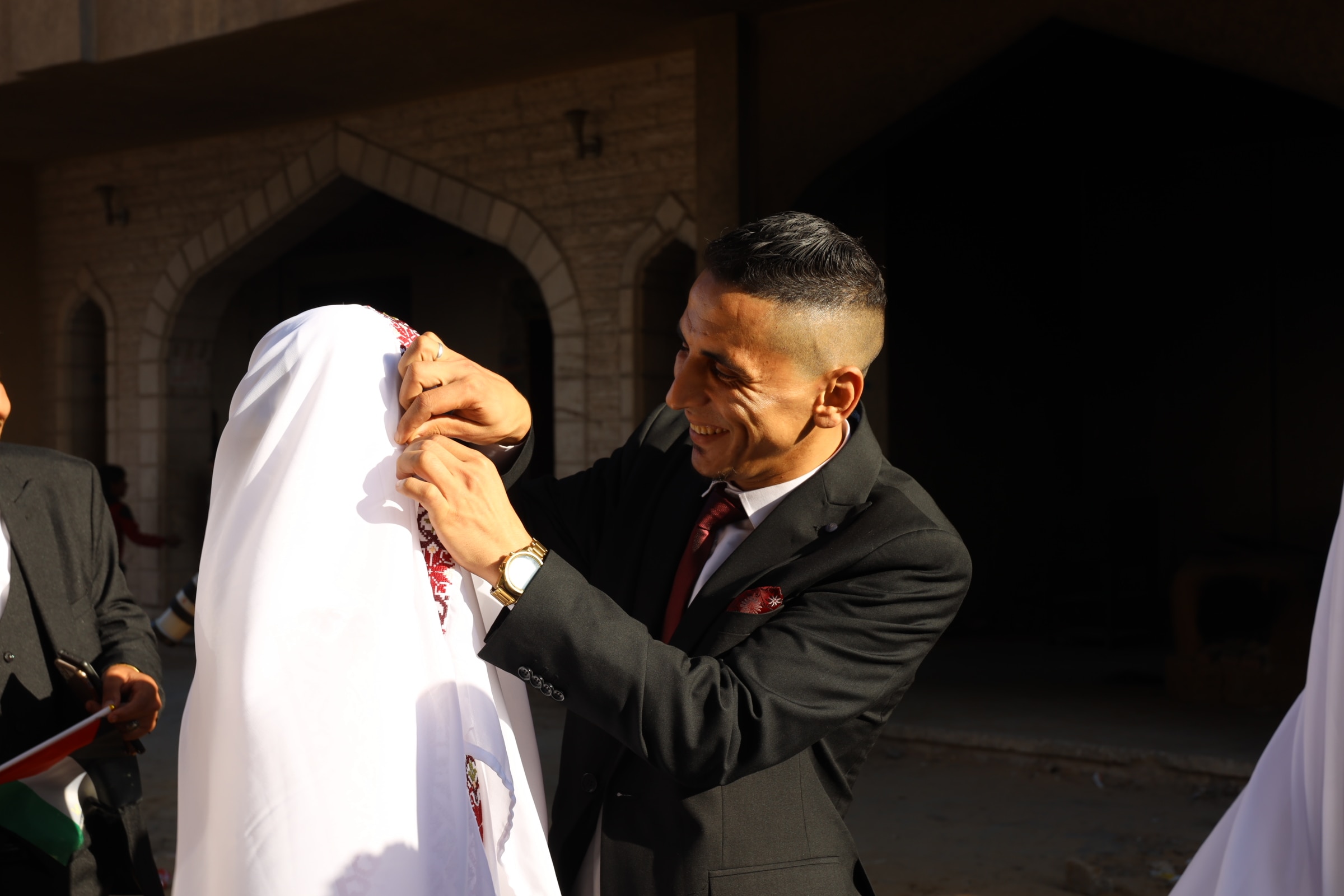A man wearing a dark suit and smiling while fixing the head scarf of a woman in front of him, outside.