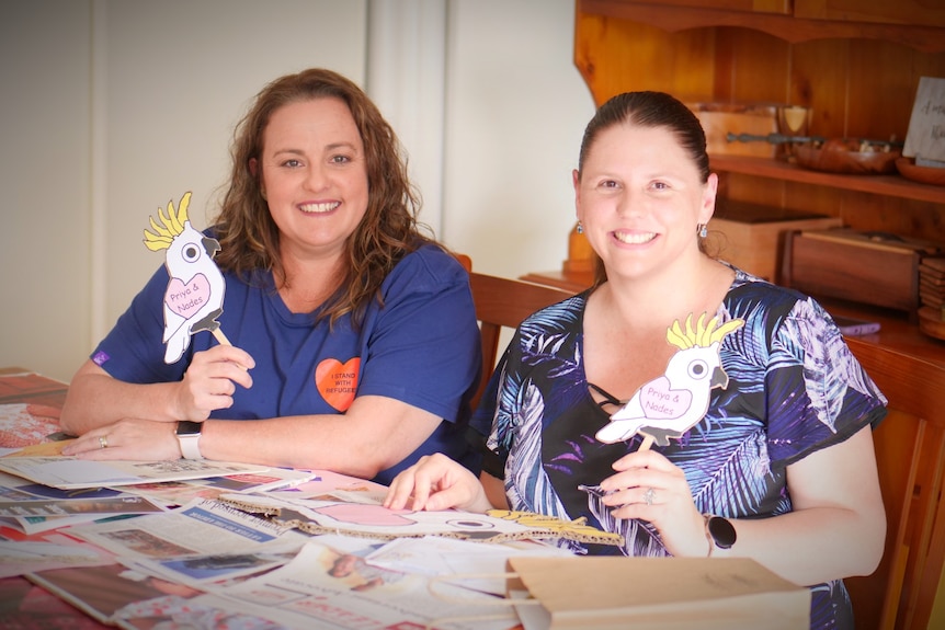 Two women sitting at a table covered in paper materials, holding up two miniature cockatoos and smiling