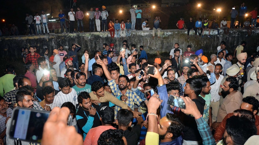 A crowd of people chant and take pictures on their smartphones at the site of a fatal rail crash