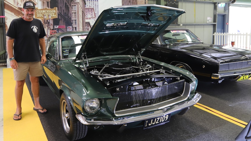 A man wearing a black shirt and cap standing next to a green Ford Mustang car, which is on display.