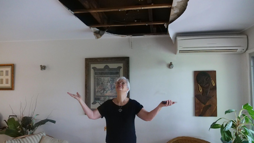 A lady with a black shirt and white pants standing under a hole in her ceiling.