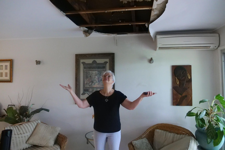 A lady with a black shirt and white pants standing under a hole in her ceiling.