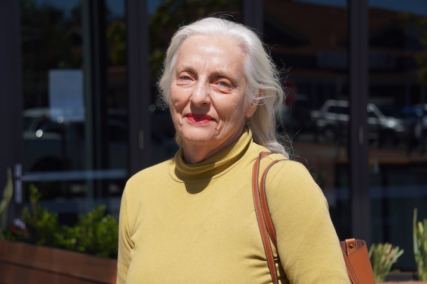 An older woman with white hair smiles at the camera wearing a yellow turtle neck top