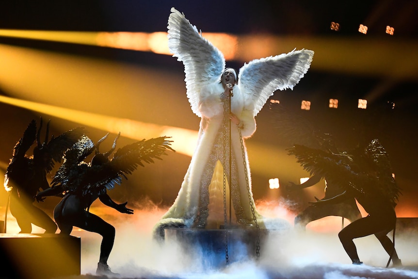 Performer Tix stands on a podium surrounded by backup dancers wearing massive white feathered wings singing into a mic