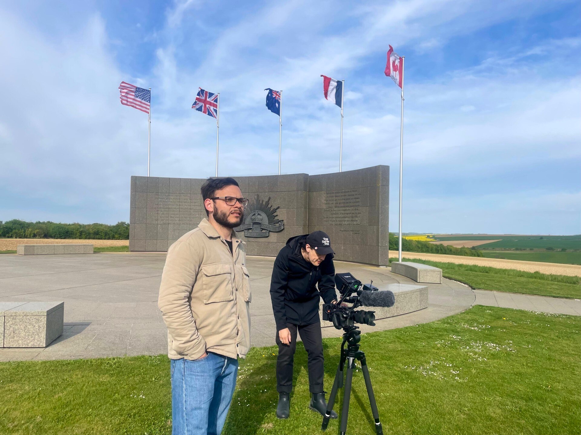 Two men stand with film camera in front of War memorial. Six countries flags fly above them in a blue sky. 