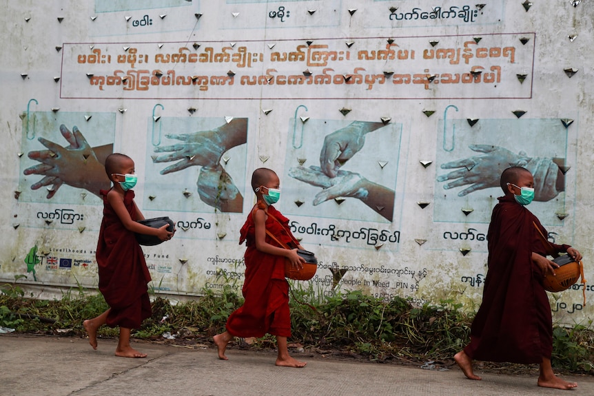 Three child monks in red robes walk past a wall showing hand-washing, wearing masks.