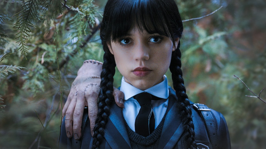 The severed hand 'Thing' sits on the shoulder of Wednesday Addams, who has long black plaits.