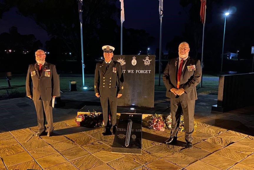 three men in uniform stand at a war memorial with flags behind them