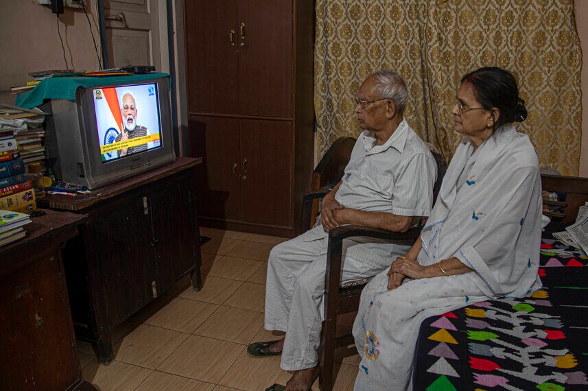 An elderly Indian couple sit in a dimly-lit bedroom and watch PM Narenda Modi on a small square TV.