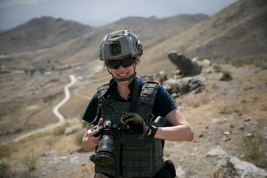Heanue wearing helmet and protective clothing holding camera standing in front of bare hills.