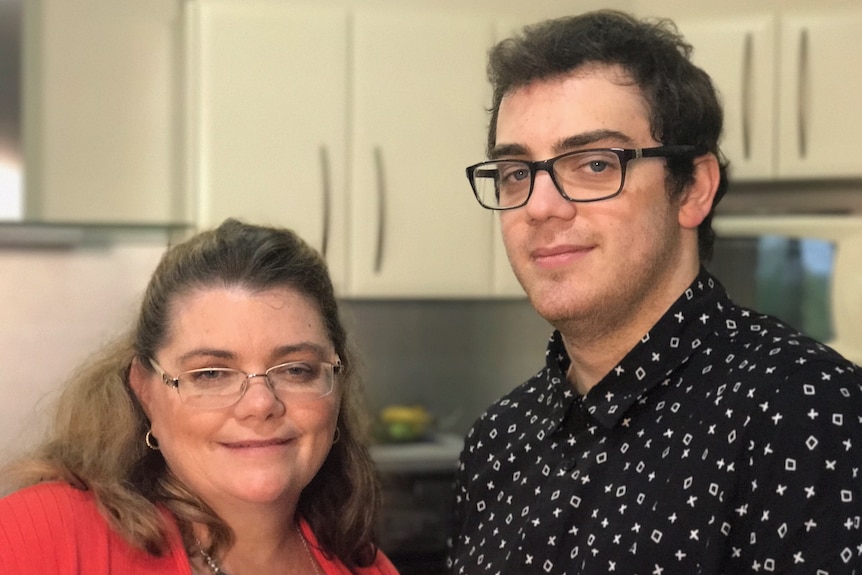 A woman in her mid-40s wears a red shirt and stands with her twenty-something yo son in their kitchen.